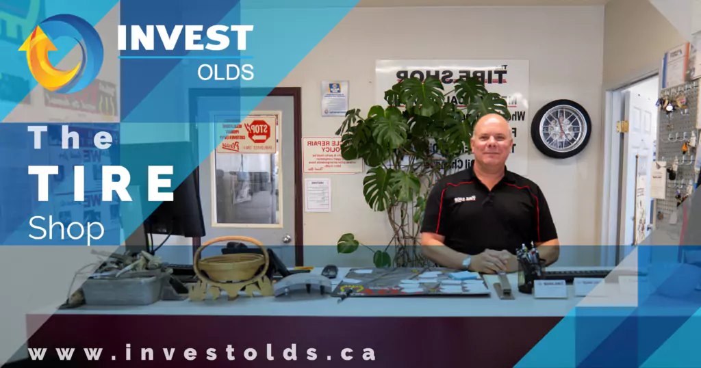 A friendly tire shop owner posing for a photo, with shelves of tires and shop equipment visible in the background.