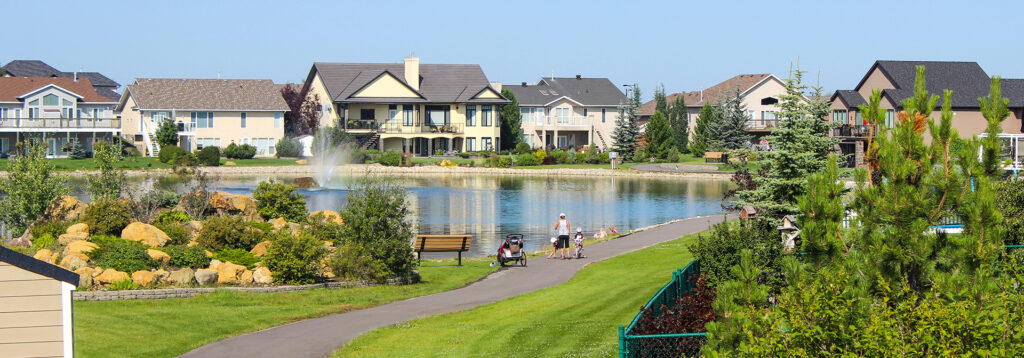 Families and individuals strolling along paths by the community pond in Olds, with residential buildings framing the serene backdrop.